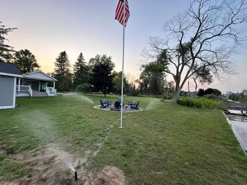 Flagpole with American flag beside a cozy lakeside yard, featuring a fire pit and seating area.