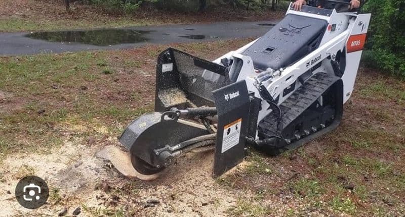 Compact track loader using a stump grinder attachment on a grassy landscape.