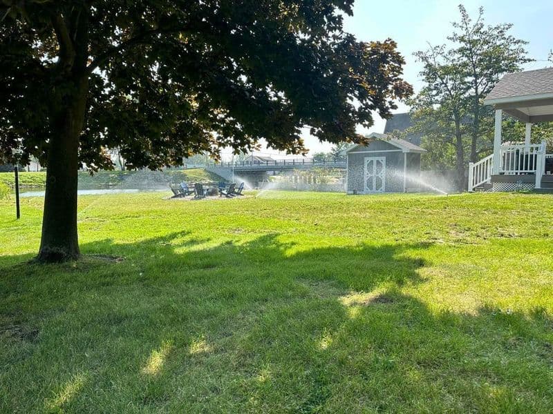 Lush green lawn with trees and patio furniture, surrounded by gentle sprinkler mist.