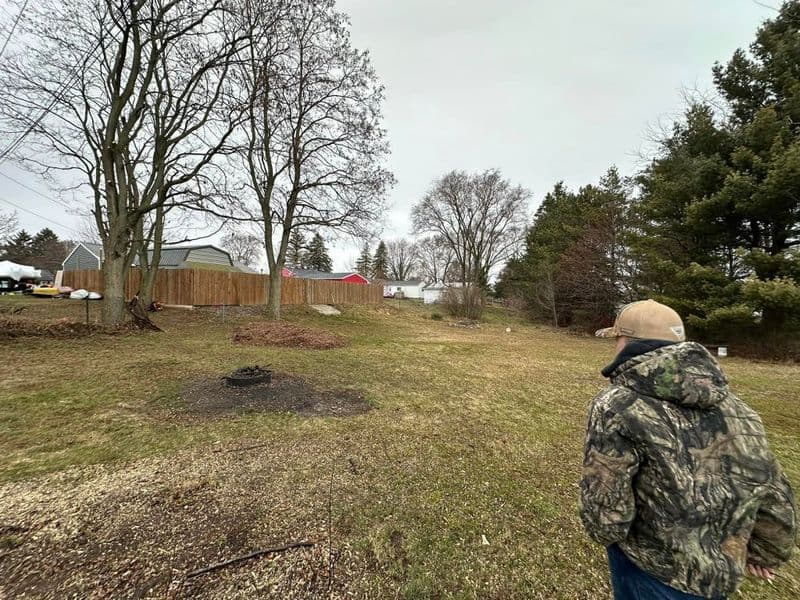 Person in camouflage jacket stands in a grassy area with trees and a rustic landscape.