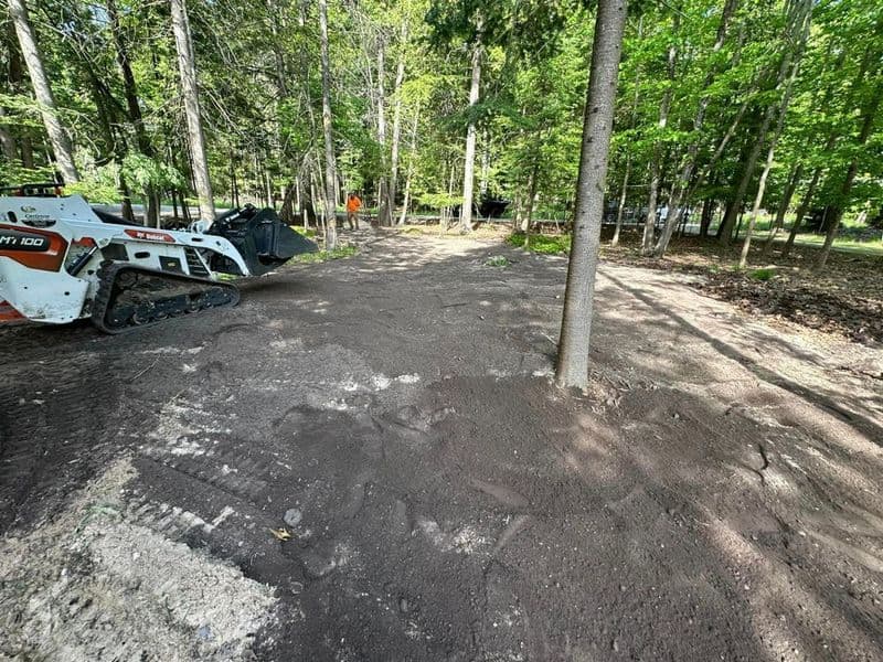 Bobcat excavator leveling ground in wooded area with tree and construction materials.