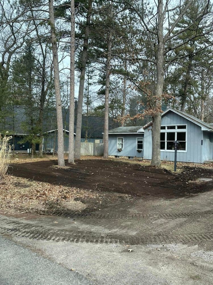 Blue cottage surrounded by trees, newly landscaped yard with freshly turned soil.