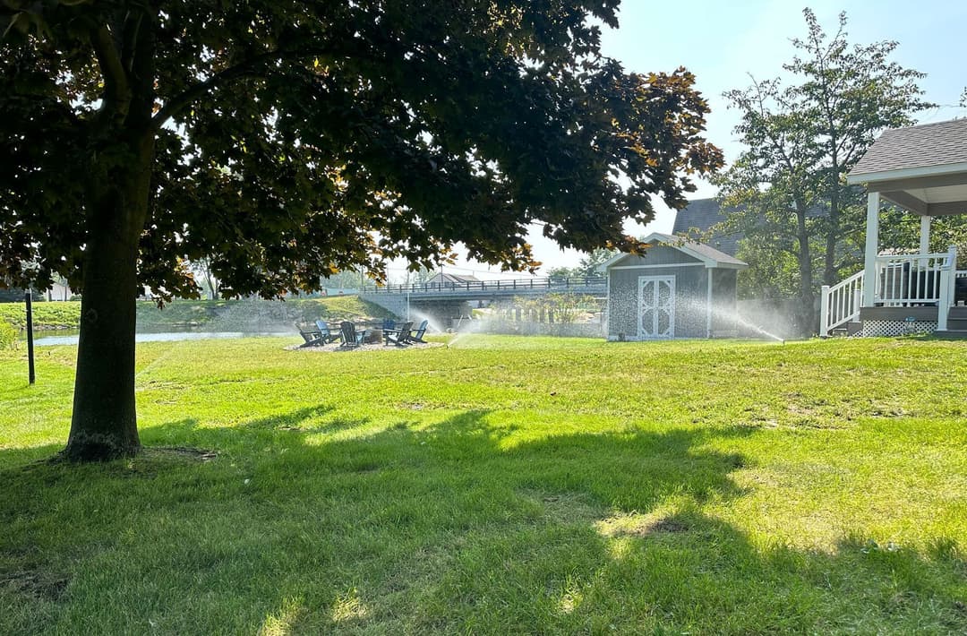 Lawn sprinkler watering grass near chairs and a shed under a sunny sky.