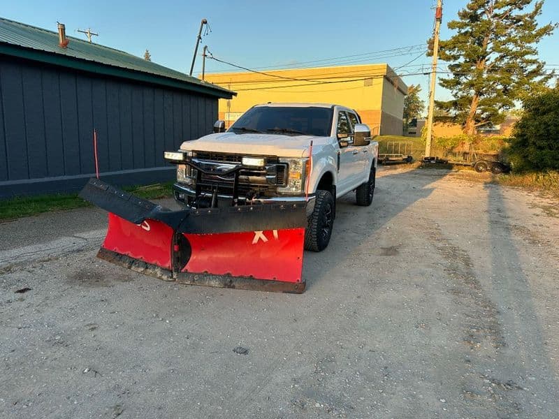 Pickup truck with a snow plow attachment parked on a dirt road near buildings and trees.
