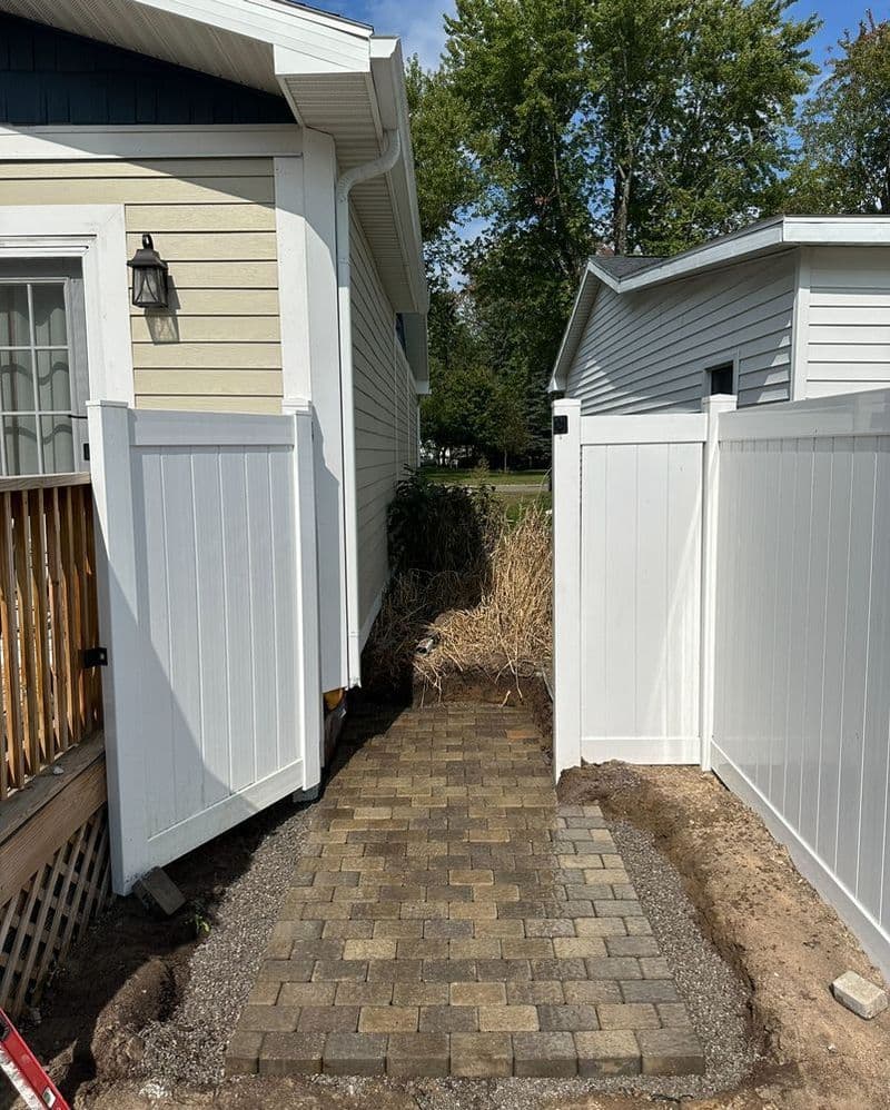 Paved walkway between two white fences leading to a side yard with trees.