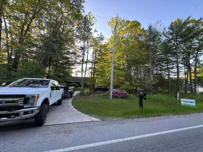 White pickup truck parked at a forested property entrance with lush greenery and a "Clean Cut" sign.
