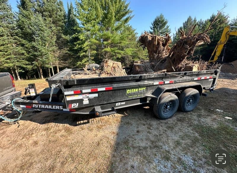 Black PJ Trailers dump trailer filled with tree debris parked in a wooded area.