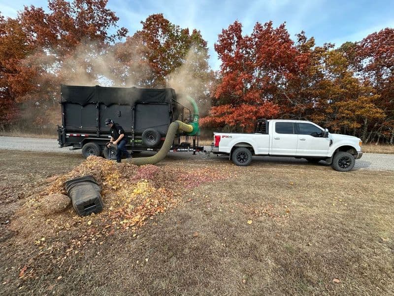 Truck with leaf vacuum collecting autumn leaves on gravel road surrounded by colorful trees.