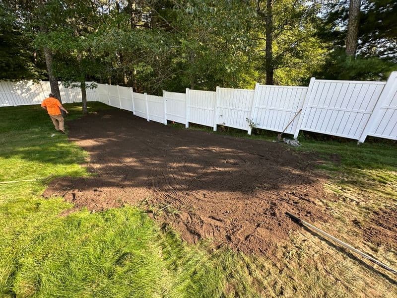 Worker preparing landscaped area with freshly tilled soil beside white picket fence.