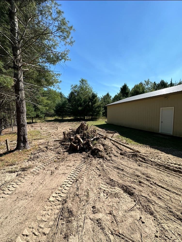Cleared area with a dirt mound and trees beside a yellow building under a clear blue sky.
